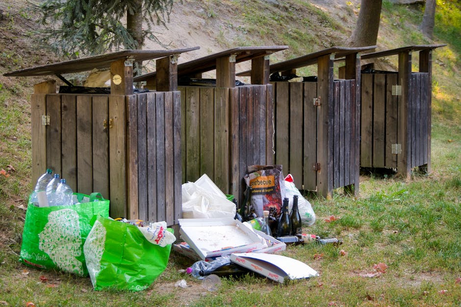 A row of four wooden outdoor rubbish bins with hinged flat roofs, positioned on a grassy area beside a sloped, wooded landscape. The bins are weathered with dark brown and blackish wood finishes, arranged side by side, each featuring metal hinges and latches. In front of the bins, there is a collection of discarded waste including a large green plastic bag containing clear plastic bottles, a partially opened white cardboard pizza box with some remains inside, empty glass bottles, a bag labeled 'sarbecute', and various smaller plastic and paper items scattered on the ground. The setting is outdoors with trees and greenery visible in the background, suggesting an informal rubbish disposal area. The scene reflects a situation where waste has been left outside for collection or disposal, illustrating the importance of proper rubbish removal services such as those offered by Rubbish Clearance Catford, particularly within the context of private waste handling or on-site clearance for properties.