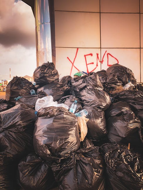 A large pile of black rubbish bags, some partially torn, is stacked against the side of a modern building with beige tiled walls. The bags are bulging with mixed waste, and several plastic bottles and other waste items are visible protruding from or resting on top of the bags. Behind the pile, a section of the building's exterior wall features graffiti that reads 'XEND' in red spray paint. The scene takes place outdoors during daytime, with a partly cloudy sky visible in the background. The surrounding environment appears urban, with some distant cityscape elements faintly visible beyond the building. The image highlights a typical instance of unmanaged waste accumulation that could relate to independent waste removal or illegal dumping, which companies like Rubbish Clearance Catford might address through professional rubbish collection services.