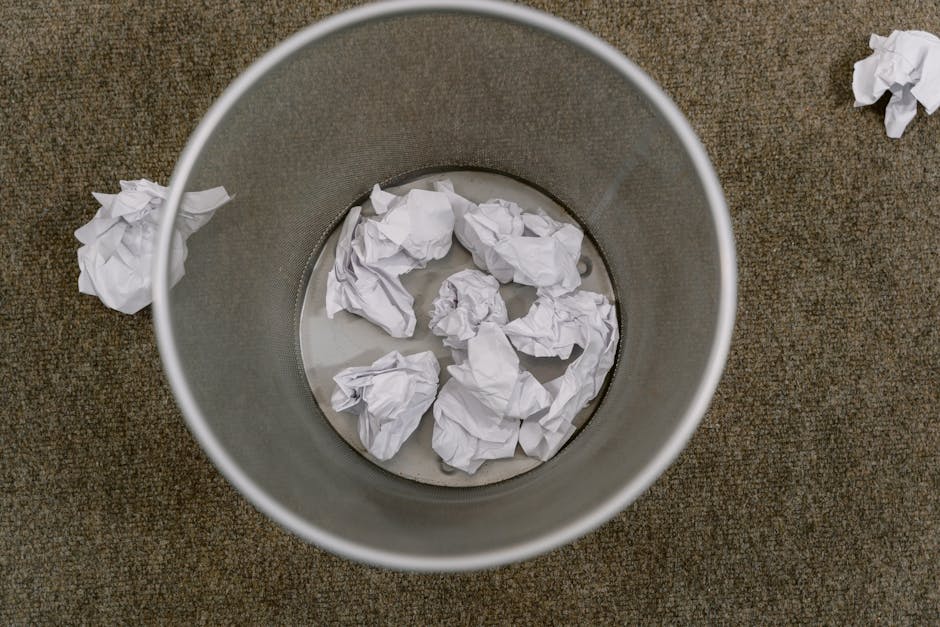 A round, metallic waste bin with a smooth, matte finish, positioned upright on a textured, brown carpeted floor. Inside the bin are several crumpled white sheets of paper, some partially submerged in a small amount of water at the bottom. Outside the bin, to the upper right, there is an additional crumpled piece of paper resting on the carpet. The scene suggests an indoor environment where waste, including paper clutter, has been collected for removal. The image's lighting is even, with no shadows or glare, emphasizing the utilitarian aspect of the waste collection process. This visual relates to private waste handling and rubbish disposal, as provided by services such as Rubbish Clearance Catford, illustrating the type of clutter often managed through independent collection or on-site clearance to avoid hidden rubbish charges and ensure proper disposal methods.