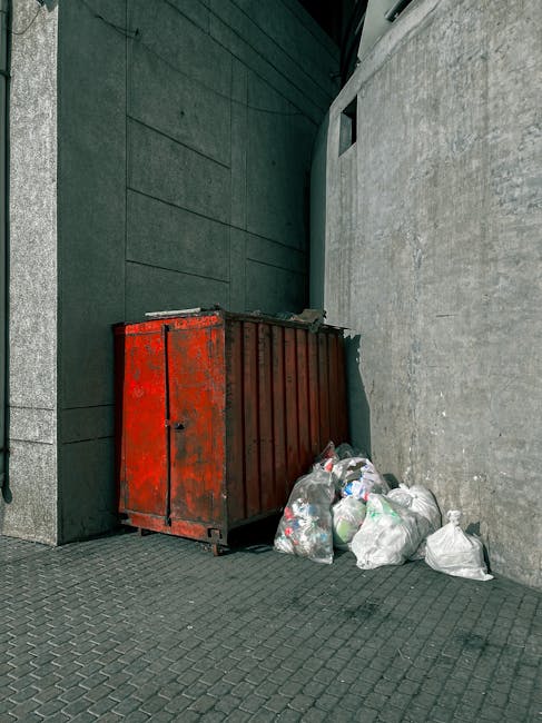 A large pile of black rubbish bags, some partially torn, is stacked against the side of a modern building with beige tiled walls. The bags are bulging with mixed waste, and several plastic bottles and other waste items are visible protruding from or resting on top of the bags. Behind the pile, a section of the building's exterior wall features graffiti that reads 'XEND' in red spray paint. The scene takes place outdoors during daytime, with a partly cloudy sky visible in the background. The surrounding environment appears urban, with some distant cityscape elements faintly visible beyond the building. The image highlights a typical instance of unmanaged waste accumulation that could relate to independent waste removal or illegal dumping, which companies like Rubbish Clearance Catford might address through professional rubbish collection services.