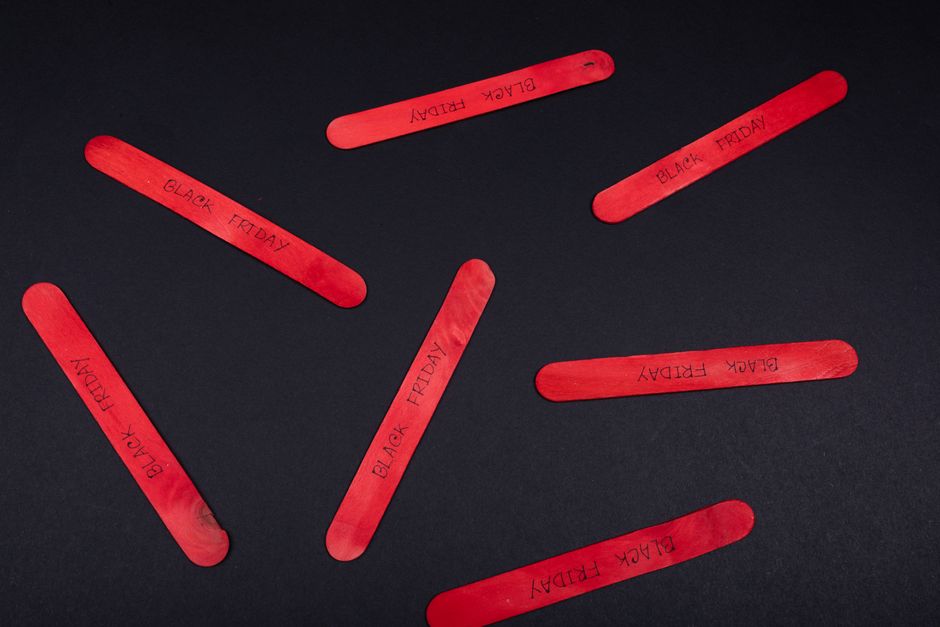 Seven red plastic foam triads with the words 'BLACK FRIDAY' written in black ink are arranged on a black surface in a scattered pattern. The triads are elongated, flat, and have rounded edges, with some overlapping slightly. The surface appears smooth and matte, providing a contrast to the vibrant red color of the foam pieces. The background is plain, emphasizing the triads' bright hue and textual markings. This image visually suggests the concept of promotional sale days, which can be associated with retail advertising, but in the context of rubbish removal services, they could metaphorically symbolize the collection of large or special waste items during specific periods, possibly indicating alternative or scheduled waste collection options. The scene's simplicity and clarity focus on the foam triads, with no other objects or environmental elements present.