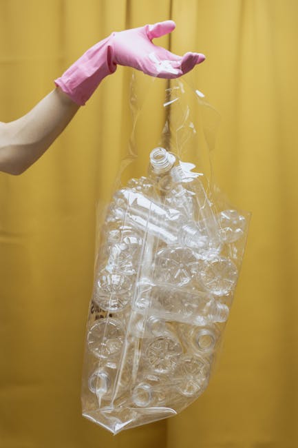 A person wearing a pink rubber glove is holding a transparent plastic bag filled with crushed, clear plastic bottles and various plastic debris. The bag appears partially filled and is suspended in front of a plain yellow backdrop, with the person's arm extended from the left side of the image. The environment suggests an indoor space used for waste handling or packaging. The plastic bottles inside the bag show visible textures of crumpled and flattened surfaces, with some caps still attached. The scene emphasizes waste collection or recycling processes, highlighting the importance of separate collection for plastics as part of private waste disposal services that may be offered by companies like Rubbish Clearance Catford. The overall composition focuses on the plastic waste container, conveying a neutral, factual view relevant to rubbish removal and environmental management without promotional language.