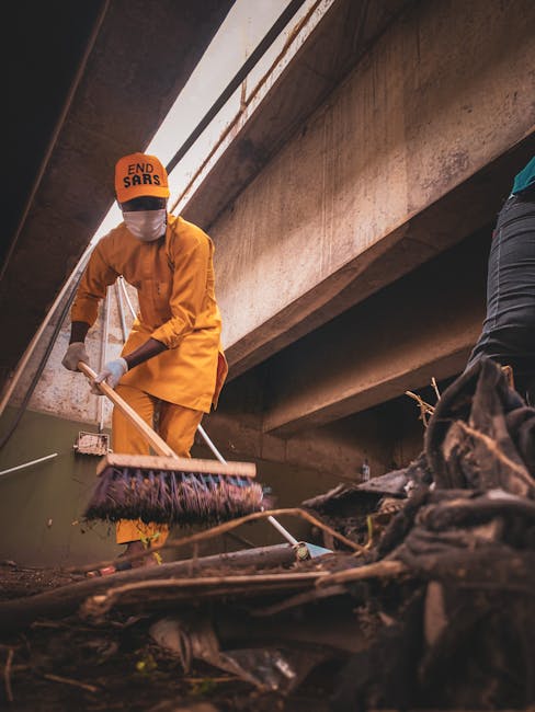 A person dressed in a yellow raincoat, gloves, and a white face mask with an orange cap displaying the words 'END SARS' is seen cleaning a waterway or drainage area beneath a large concrete bridge. They are using a long-handled brush or tool to remove debris and litter from the muddy water, which contains scattered rubbish including plastic and organic waste. The concrete bridge above has visible structural beams and a weathered surface, with sunlight filtering through gaps, casting a warm glow on the scene. To the right, part of another person is visible, wearing dark clothing and standing on the muddy bank. This scene relates to waste management and cleaning efforts that could be part of independent rubbish clearance or environmental maintenance services, such as those provided by Rubbish Clearance Catford, emphasizing the importance of on-site waste removal and environmental health.