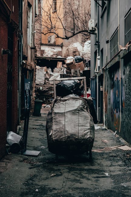 A narrow urban alleyway cluttered with accumulated rubbish and discarded materials, with a large black and gray wheeled rubbish bag prominently positioned in the foreground. The alley is surrounded by weathered brick and metal walls that feature graffiti and peeling paint, indicating a neglected environment. Behind the bag, there is an extensive pile of cardboard boxes, plastic waste, and other debris stacked against a wooden barrier or fence at the rear, suggesting on-site waste accumulation for rubbish removal. To the right, a metal building with grated windows and pipes extends along the alley, while on the left side, a partially visible door and a small trash bin can be seen. The ground is uneven, with patches of dirt and grime, and the lighting is muted, highlighting the dusty, cluttered scene. This setting reflects an area where private waste disposal or alternative rubbish collection might be necessary, as supported by a local service such as Rubbish Clearance Catford.
